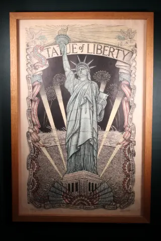 Full frontal view of James Grashow's 1986 Statue of Liberty woodcut, featuring Lady Liberty surrounded by American flags, fireworks, radiating light, decorative banners, and intricate historical imagery. Presented vertically in a wooden frame.